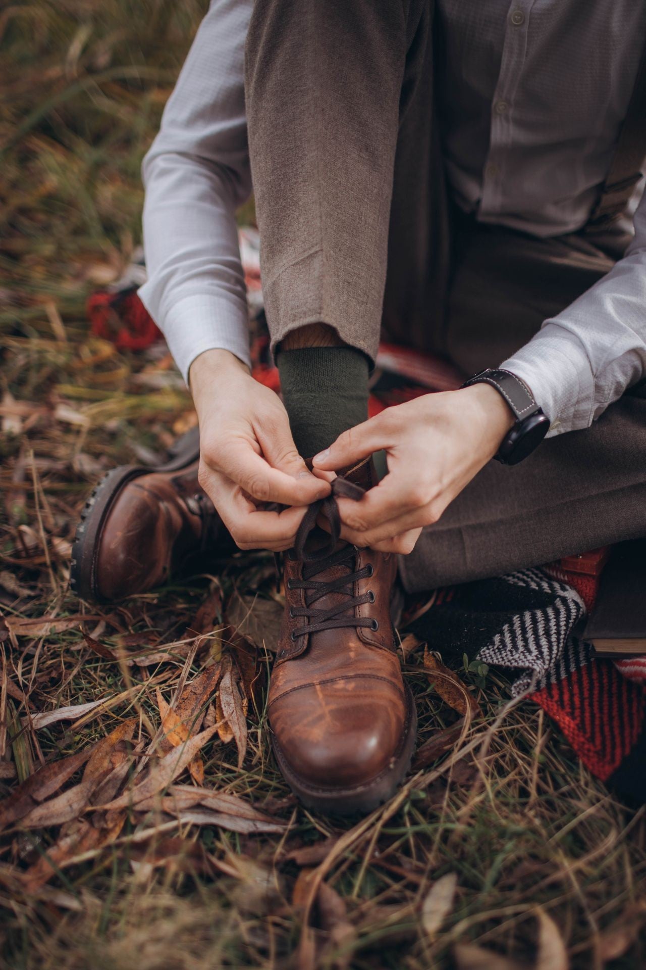 Person tying brown boots in a natural setting with grass and leaves.